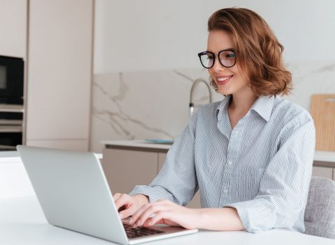 elegant-smiling-woman-in-glasses-and-striped-shirt-using-laptop-computer-while-siting-at-table-in-kitchen (1)